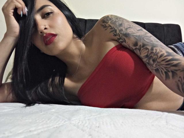 GiulianaFetich, laying on a bed, wearing a red tube top, showing her tattooed arm, and red lips, with long black hair, and a subtle smile, looking at the camera.