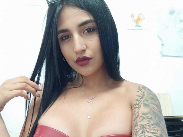  GiulianaFetich with long black hair, red lipstick, nose ring, small heart necklace, tattooed arm, holding hair, wearing red strapless top, showing her tits, white background.