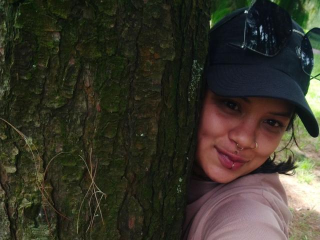 MariGonz con il piercing al naso e la lingua fuori, appoggiata a un albero, indossa un berretto nero con gli occhiali da sole sopra, una camicia rosa, mostrando il suo viso carino..
