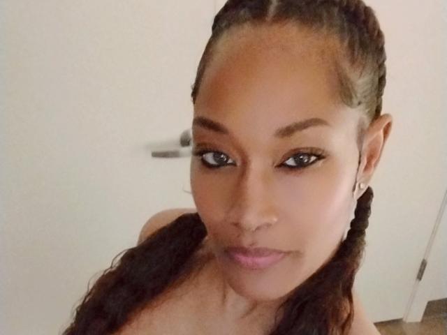 CarmelDelightF with medium brown skin, braided hair, and pink lipstick, looking up with black eyeliner, small nose ring, and earrings, showing her bare shoulders and a slight smile.