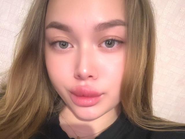 NanetteTrixie's face, showing light brown hair, green eyes, and pink lips with a slight pout, wearing a black shirt, against a white textured wall.