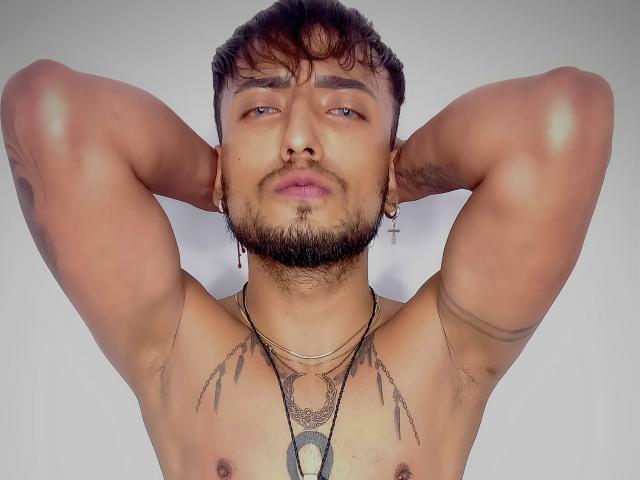 FerlucyGuy69, shirtless with arms behind head, brown skin, short dark hair, beard, tattoos on chest, necklace, earrings, looking at camera with slight smirk, blue eyes, relaxed.