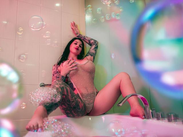 KiraKane, sensuelle dans une salle de bain, portant une combinaison transparente et scintillante, assise dans une baignoire avec sa main sur ses seins, les jambes écartées, entourée de bulles, avec des tatouages sur le bras et la jambe..