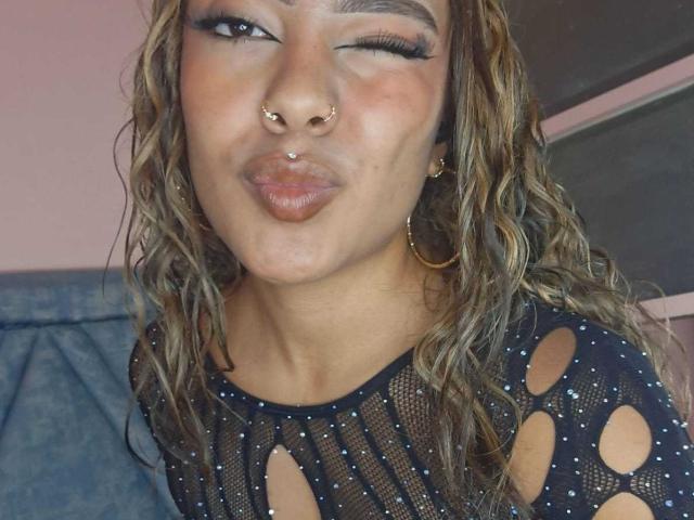 AmelieDuvall with wavy brown hair, nose ring, and hoop earrings, making a pouty face, wearing a black mesh top with cutouts, showing her tits.