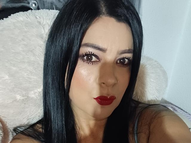 MelannySexy has shiny black hair, red lipstick, and brown eyes with long lashes, pouting her glossy red lips. She's looking directly at the camera with a slightly surprised expression.