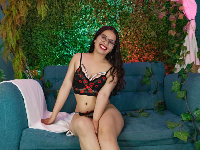 Julia Waine, avec des lunettes, du rouge à lèvres et de la lingerie en dentelle noire ornée de motifs de roses rouges, est assise sur un canapé bleu turquoise, les jambes jointes, avec un tissu blanc à sa gauche, et elle sourit..