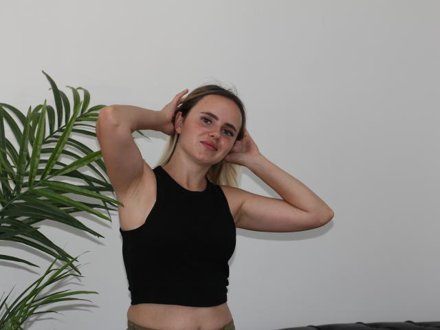 MilanaBloom with arms up, black sleeveless top, blonde hair, smiling, small tits, pale skin, green plant on left, white wall, indoor setting, casual pose, relaxed expression.