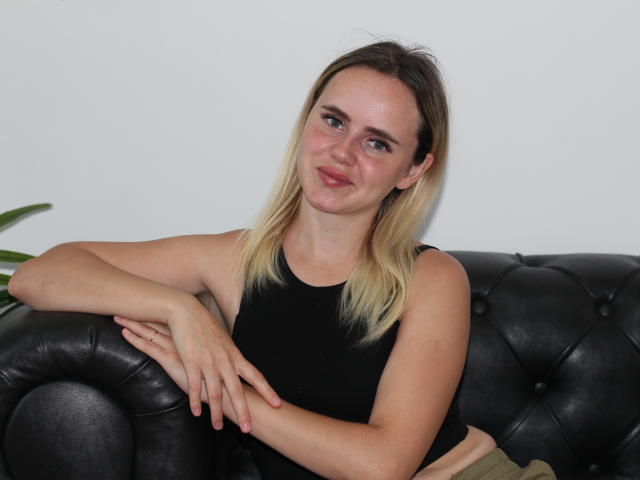 MilanaBloom with blonde hair, black tank top, and tan pants, sitting on a black leather couch, arms resting on top, smiling slightly, looking relaxed.