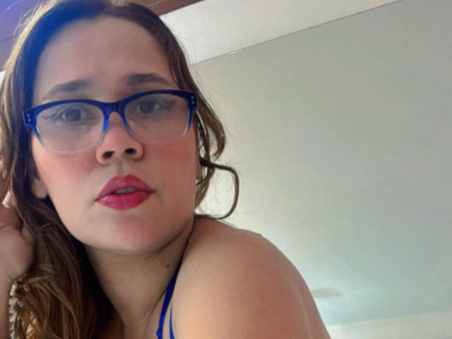 SophiaQueens mit blauen Brillen, rotem Lippenstift und welligem braunem Haar, die ihre freie Schulter zeigt und einen leichten Hauch von Röte im Gesicht hat. Ihre Brüste sind teilweise sichtbar..