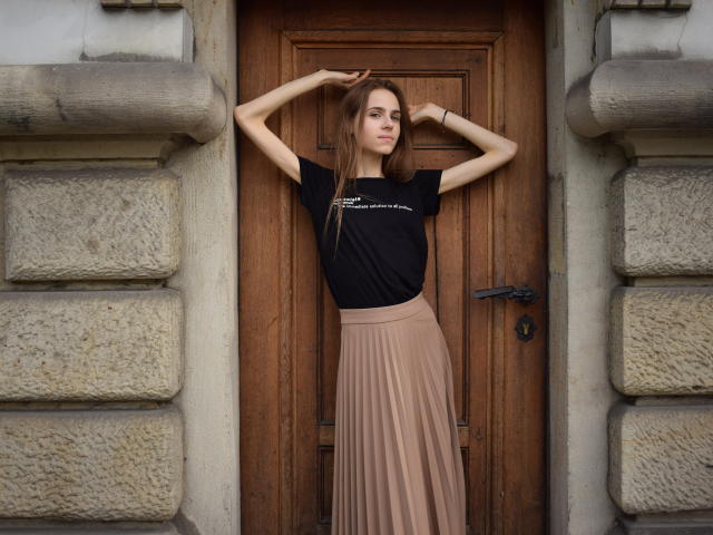 GlamKatrisse stands in front of a wooden door, arms raised, wearing a black top and a pleated beige skirt, with a slim, tall, and attractive physique.