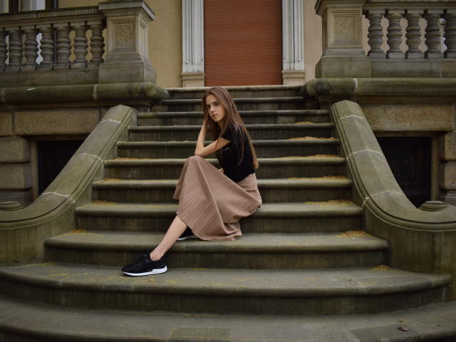 GlamKatrisse with long brown hair, black t-shirt, and beige skirt, sitting on stone stairs, black sneakers, looking down, arms crossed, butt resting on stairs, serious expression.
