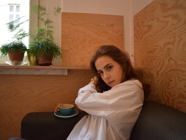 GlamKatrisse with wavy brown hair, white shirt, crossed arms, looking at camera, sitting next to green potted plants, blue cup on dark gray couch, wooden wall background.