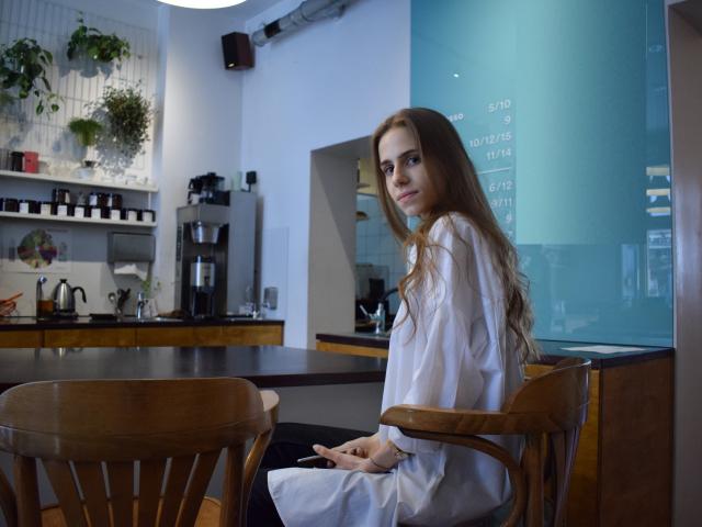 GlamKatrisse with long brown hair sits in a wooden chair, wearing a white shirt, looking over her shoulder, in a cafe with wooden tables and potted plants.
