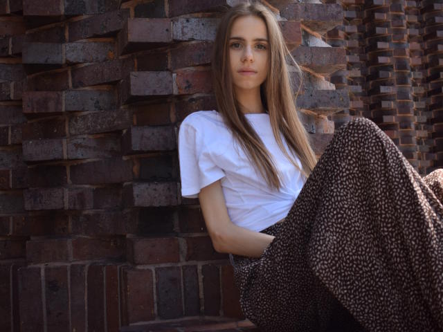GlamKatrisse with long brown hair, white tee, and brown polka-dot pants, sitting against a brick wall, knees up, looking straight at the camera, slightly serious expression.
