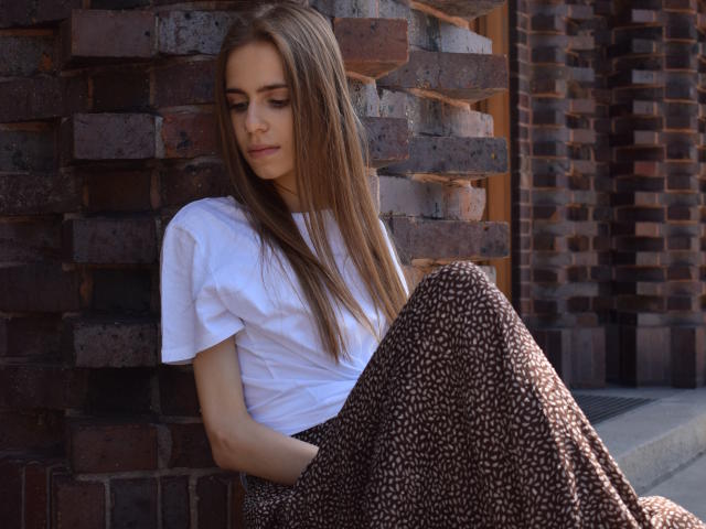 GlamKatrisse with long brown hair, white tee, and brown leopard print skirt, sitting against a brick wall, looking down with a subtle, serious expression. Hands in skirt pockets.