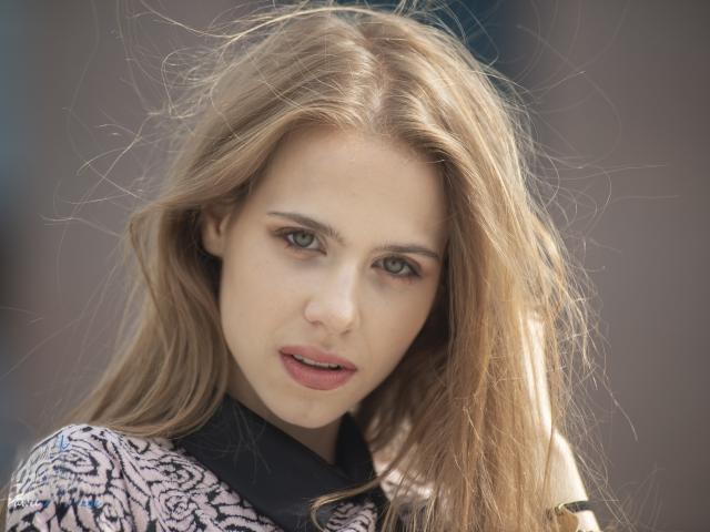 GlamKatrisse has long, messy blonde hair, green eyes, and a slight open-mouth expression. She's wearing a black and pink patterned top, looking directly at the camera.