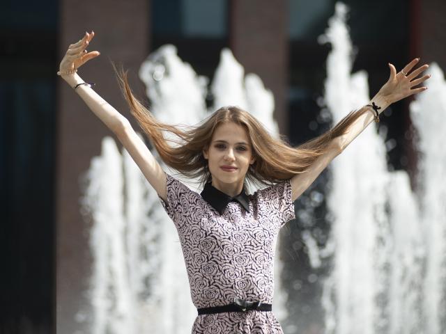 GlamKatrisse with arms raised, long brown hair flowing, wearing a pink and black patterned dress, standing in front of a white water fountain, smiling with red nails.