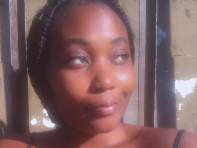 DarkGodesss has shiny brown skin, short braided hair, and a subtle smile with her eyes looking to the side. Her glossy lips are slightly parted. Soft light highlights her face.