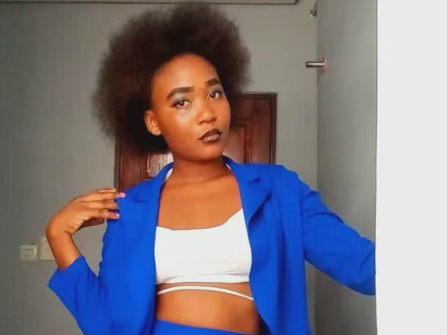DarkGodesss with dark skin, afro, wearing blue blazer, white crop top, showing midriff, hand on blazer, looking at camera, in a simple room.