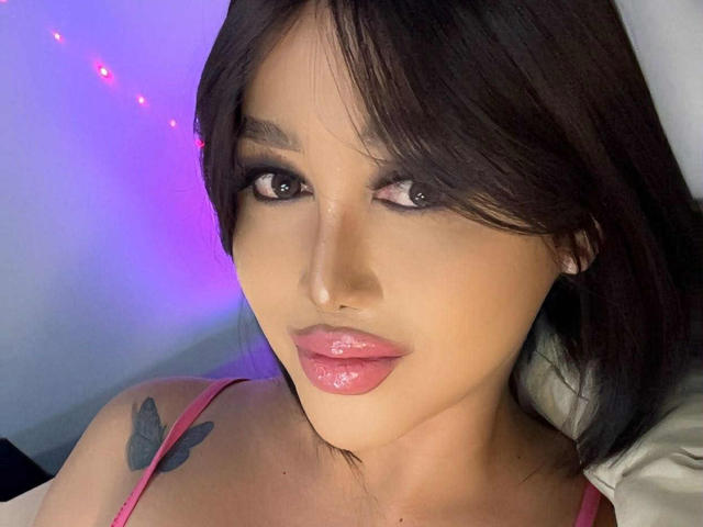 CallieStewart with glossy pink lips, dark brown hair, and a butterfly tattoo on her right shoulder. Her tits are partially visible in a pink top.