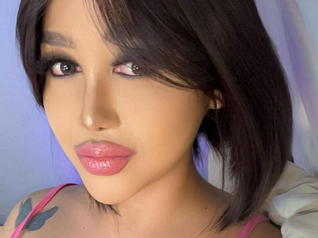 CallieStewart has glossy pink lips, dark brown eyes with smoky makeup, and a short black bob. She's wearing a pink top, showing a tattoo on her right shoulder.