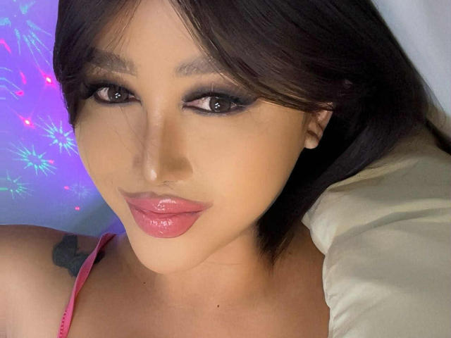 CallieStewart's face with glossy pink lips, dark makeup, and brown hair, wearing a pink top, lying on a white pillow. Her tattoo is partially visible on her shoulder.