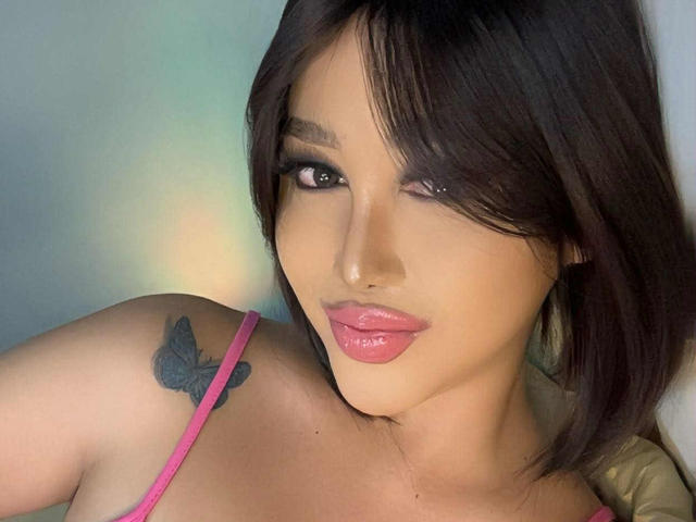 CallieStewart has pink lipstick, dark brown hair, and a butterfly tattoo on her shoulder. She's wearing a pink top with thin straps. Her eyes are slightly winking.