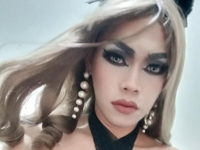 InnocentFameHottie with blonde hair, dark eye makeup, pearl earrings, black choker, and a serious expression. Fluffy hair, eyeliner, and glossy lips. Slight wind in hair.