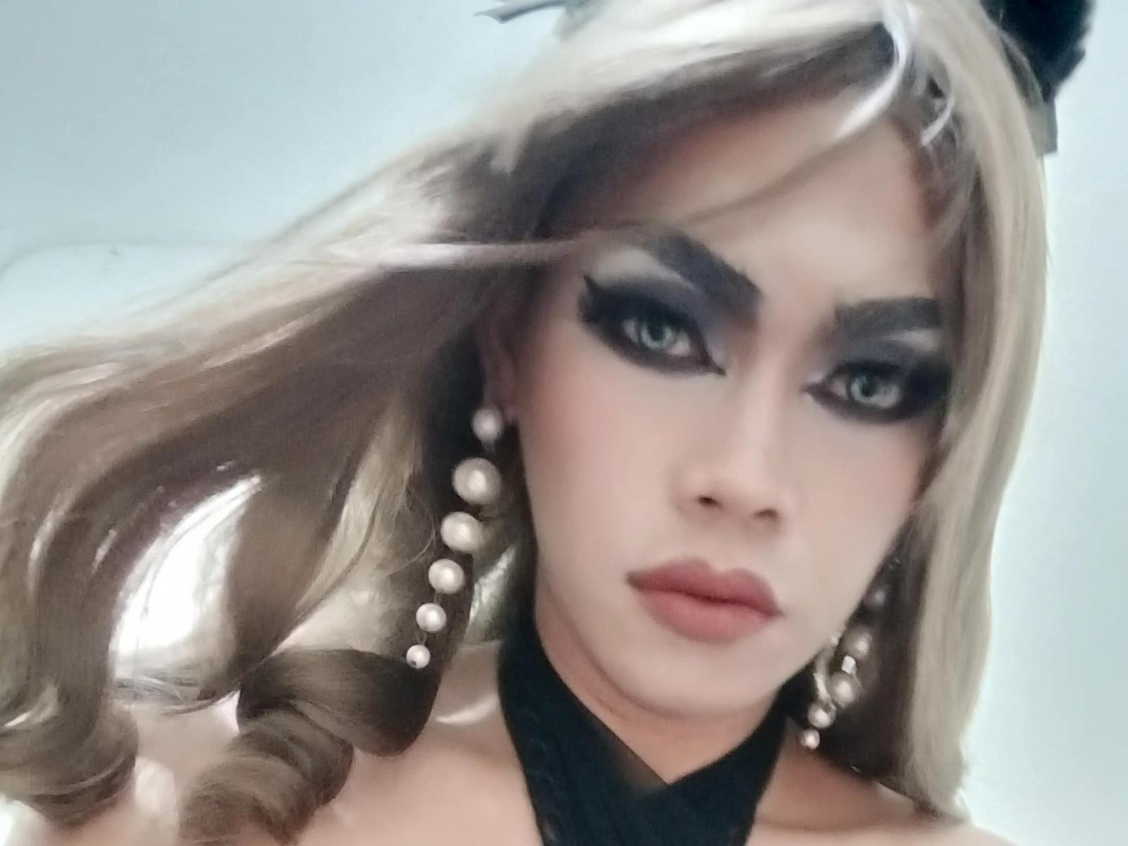InnocentFameHottie with blonde hair, dark eye makeup, pearl earrings, black choker, and a serious expression. Fluffy hair, eyeliner, and glossy lips. Slight wind in hair.
