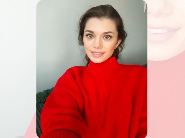 EvaMcCoy with green eyes, wearing a red turtleneck, smiling slightly, brown hair in a loose ponytail, soft light on her face. Her tits are subtle under the sweater.