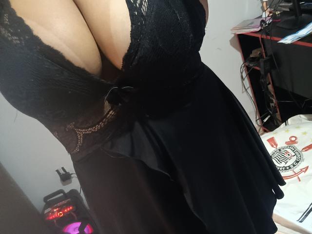 CabelereiraSexy69's large, bare boobs are barely covered by a black lace dress with a deep cleavage. Black fabric and lace details are visible.
