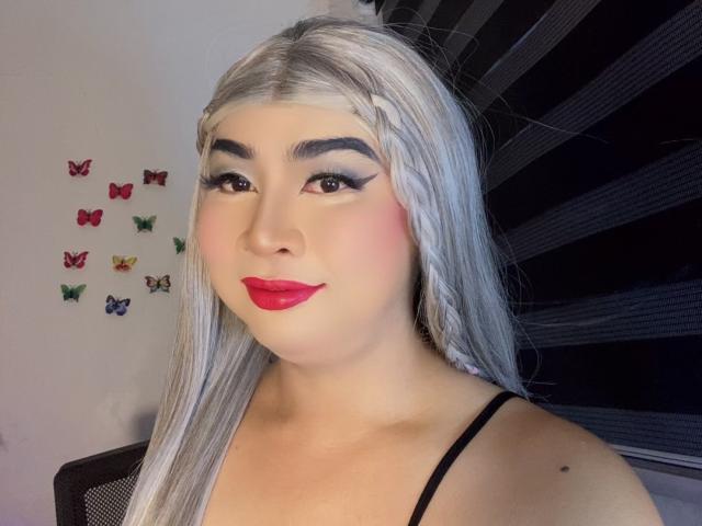 AsianFemboyX with long silver hair, braided crown, thick black eyeliner, red lipstick, smiling, wearing a black strap top, and butterfly wall decorations.