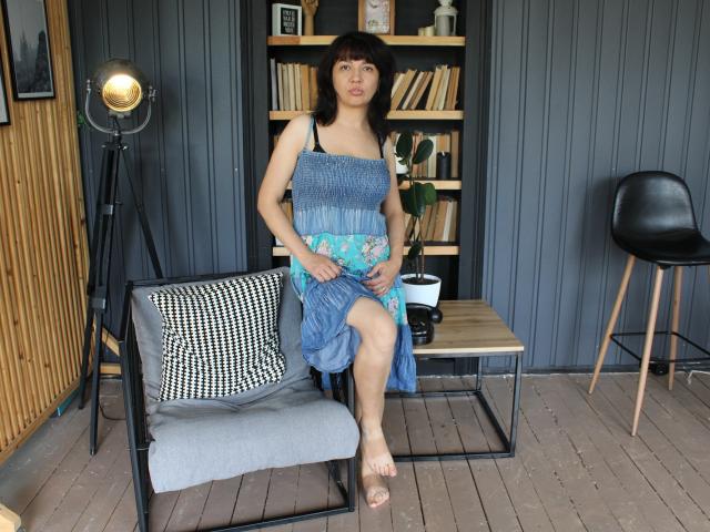 SelenaFly, Asian woman, small tits, wearing blue dress, sitting on chair, black tripod light on left, black chair on right, wooden shelf with books behind, wooden floor.