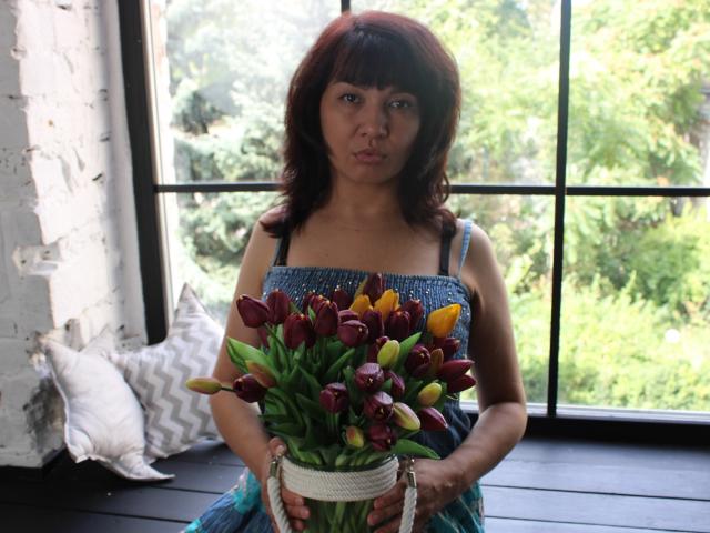 SelenaFly with red hair, blue dress, holding bouquet of red and yellow tulips, standing in front of window with green trees outside, pouty face, holding bouquet with white basket.