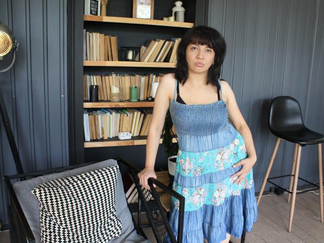 SelenaFly with medium tits, black bangs, wearing a blue floral dress, stands in a room, hand on chair, black chair on right, bookshelf behind, wooden floor.