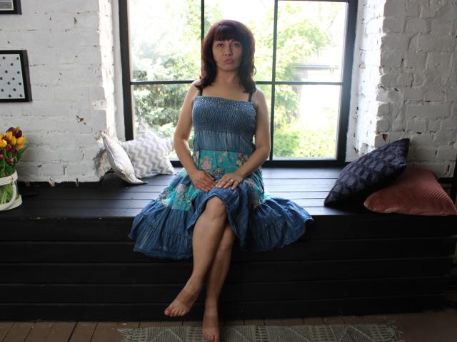 SelenaFly, barefoot, sitting on black wooden window seat, wearing blue floral dress, arms crossed, legs crossed, white brick wall, window, and pillows in background.