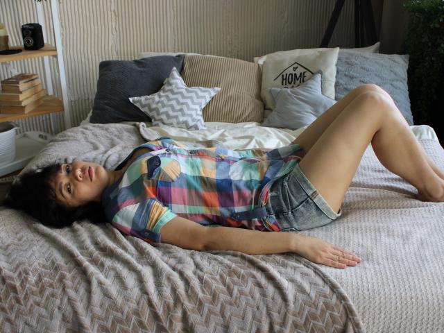 SelenaFly is lying on a bed, wearing a colorful plaid shirt and denim shorts, with her ass up and her head on the pillow, looking at the camera.