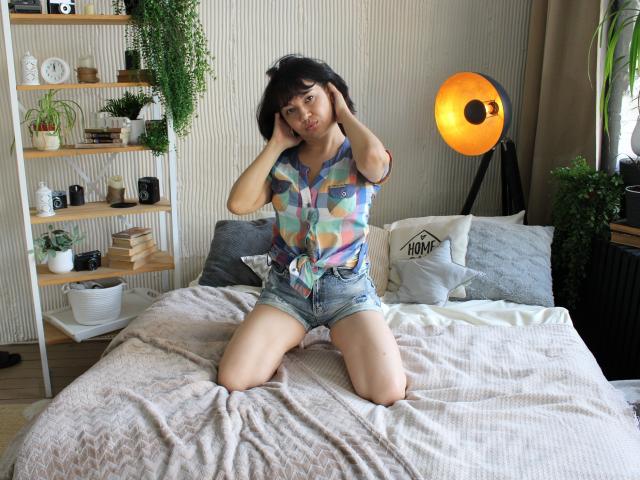 SelenaFly, with hands in her black hair, sits on a bed in colorful tie-front shirt and denim shorts, knees apart, looking at camera, with a soft expression.