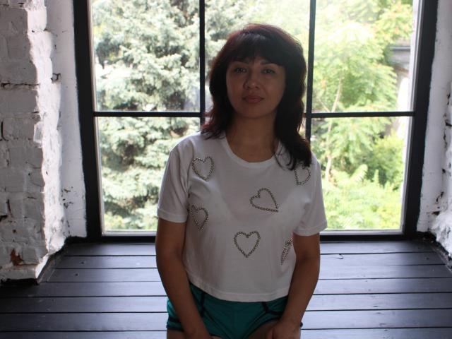 SelenaFly has medium brown hair, wearing a white shirt with heart patterns and teal shorts, standing in front of a window with green trees outside.
