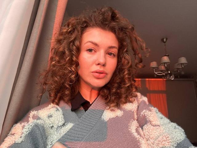 CurlyCandyX has curly brown hair, light skin, and is wearing a fluffy, patterned sweater with blue and white patches. She's looking at the camera with a pouty expression.
