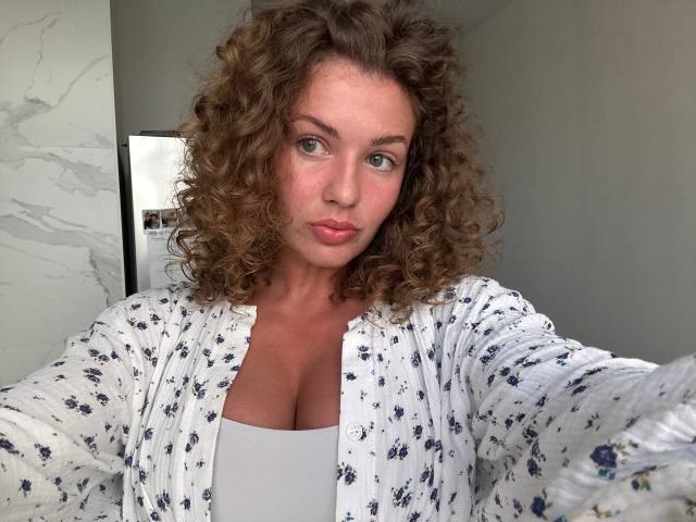 CurlyCandyX with curly brown hair, pouty pink lips, and green eyes, wearing a white floral robe, showing her large, round tits in a white top.