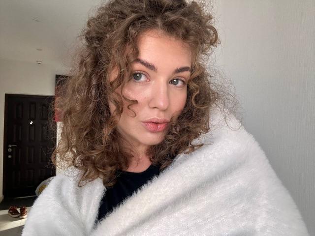 CurlyCandyX has curly brown hair, green eyes, and a cute face with pink lips, wearing a white fuzzy coat, black shirt, and a subtle, sexy look.
