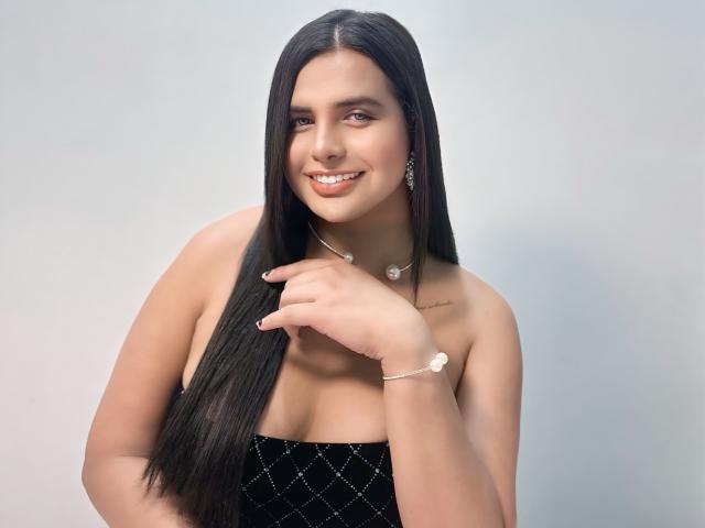 HotSelenaTrans has long black hair, tan skin, small tits, smiling with her hand near her chin. She's wearing a black strapless dress, silver earrings, and a bracelet.