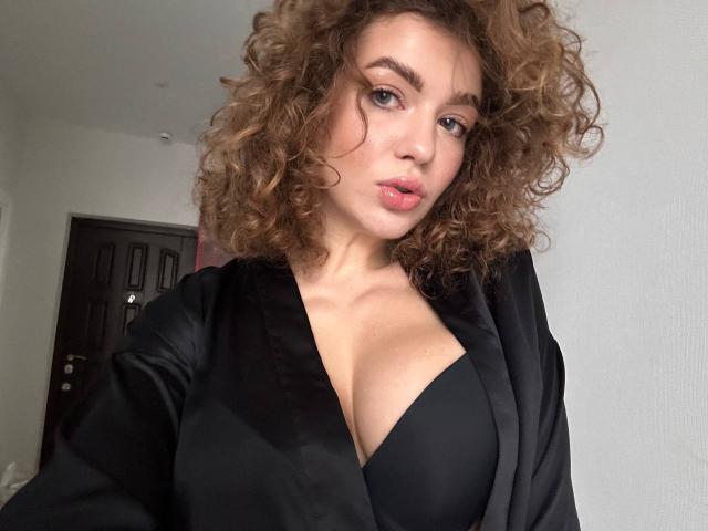CurlyCandyX with curly brown hair, pouty lips, and a black bra, wearing a black satin robe, showing her tits. She's looking at the camera with a slight smirk.