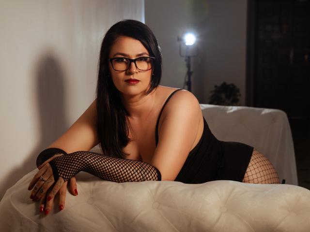 CarolinnaSaenz with black hair, glasses, red lipstick, black fishnet gloves, and black dress, leaning on white bed, showing her tits and red-painted nails. Soft light in background.
