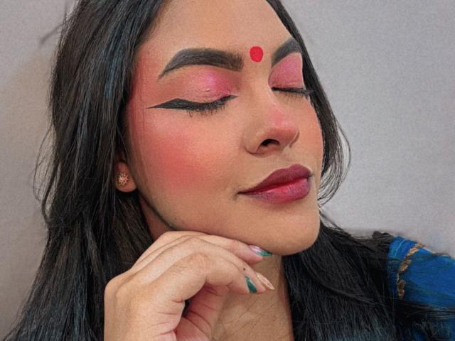 LakshmIrani with closed eyes, pink eyeshadow, black eyeliner, red dot on forehead, pink blush, red lipstick, dark hair, blue and gold patterned top, resting chin on hand.