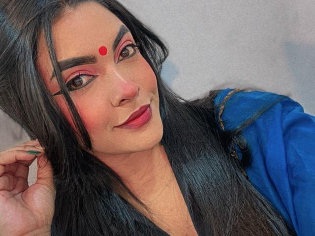 LakshmIrani with long black hair, blue top, pink eye makeup, red dot on forehead, dark lipstick, and green nails, smiling slightly, hand in hair.