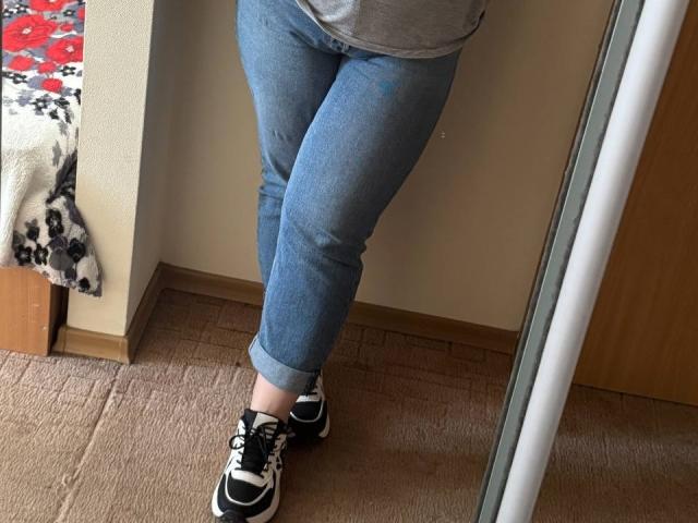 Leiliia's ass is in tight blue jeans, and she's wearing black and white sneakers with her legs crossed, standing on a carpeted floor, reflected in a mirror.