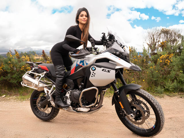SofyaLane, con lunghi capelli castani, un abito nero e stivali neri, seduta su una moto BMW bianca con dettagli neri e rossi, su una strada sterrata. Il suo sedere è visibile..