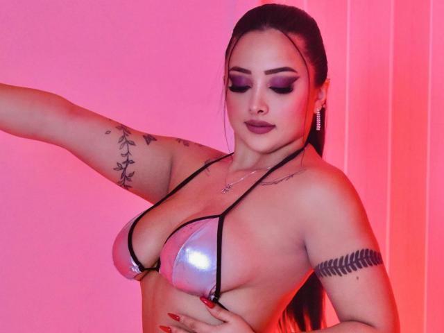 KylieCharm avec un haut de bikini argenté brillant, de grandes seins, des tatouages sur le bras et le côté, du rouge à lèvres foncé, du fard à paupières violet, un éclairage néon rose et de longs cheveux bruns attachés en queue de cheval..